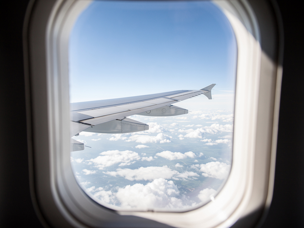 An airplane wing above soft white clouds with blue sky seen through a cabin window.