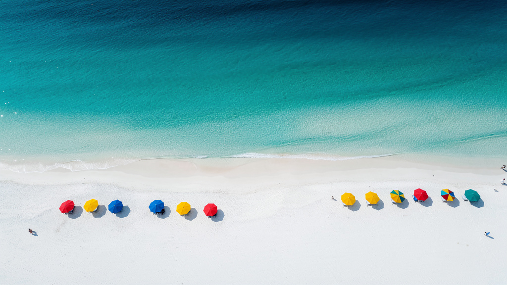 Wide aerial view of a turquoise beach with white sand and a few colorful umbrellas under bright sun.